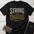 Strong and Courageous t-shirt in Black — Joshua 1:9 Christian tee flat lay with Holy Bible and ichthys on whitewashed wood