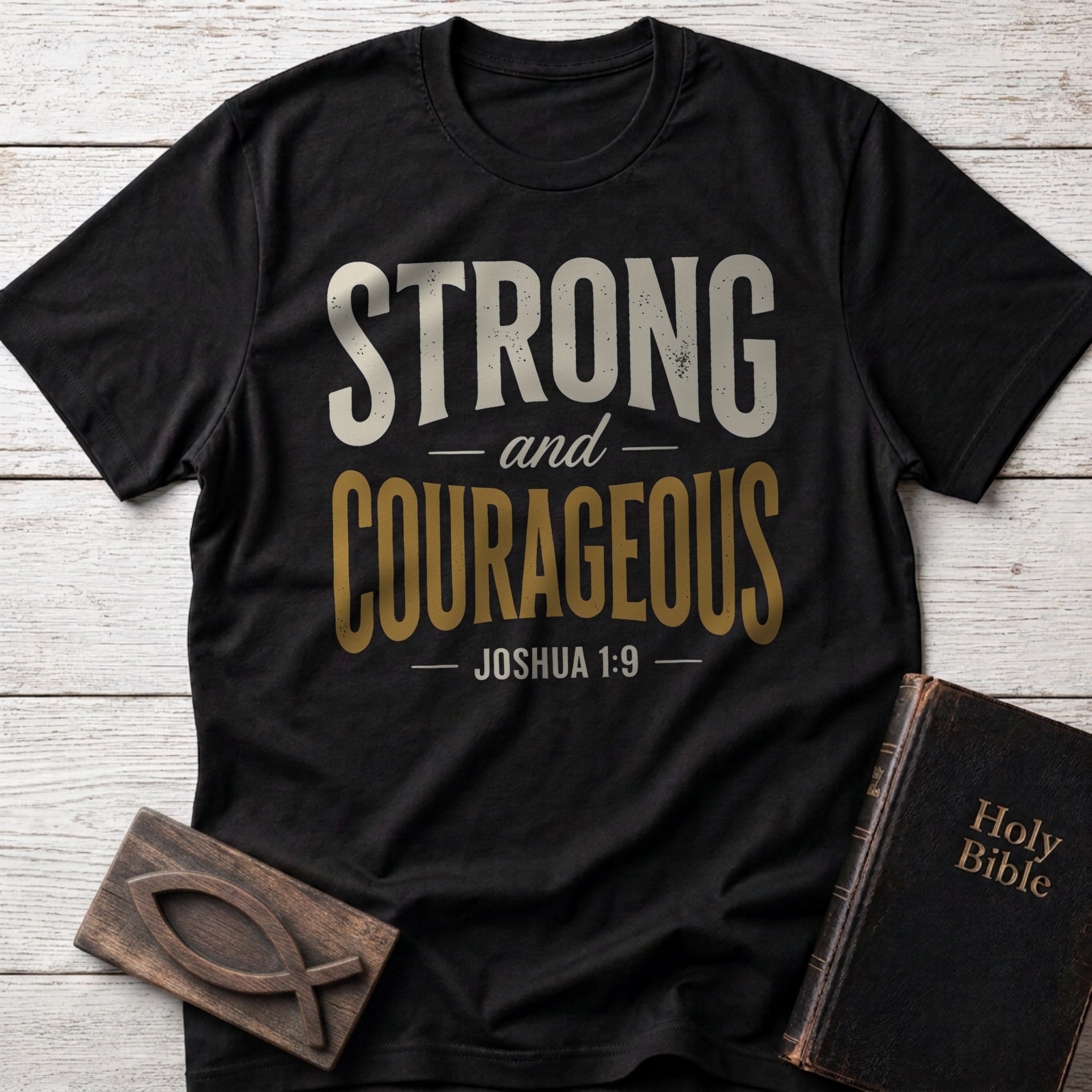Strong and Courageous t-shirt in Black — Joshua 1:9 Christian tee flat lay with Holy Bible and ichthys on whitewashed wood