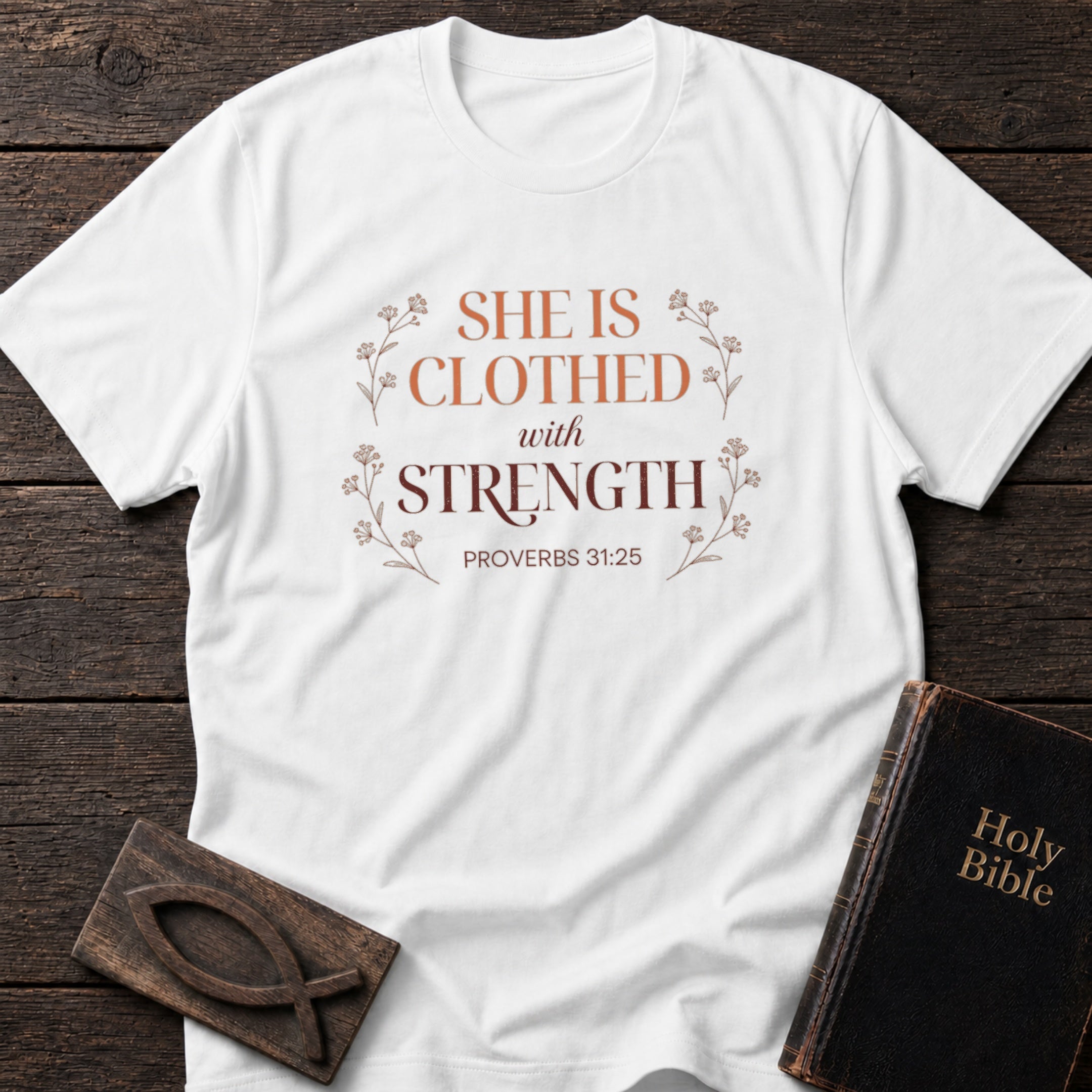 She Is Clothed With Strength t-shirt in White — Proverbs 31:25 Christian tee flat lay with Holy Bible and ichthys on dark wood