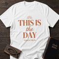This Is the Day t-shirt in White — Psalm 118:24 Christian tee flat lay with Holy Bible and ichthys on dark wood