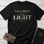 The Lord Is My Light t-shirt in Black — Psalm 27:1 Christian tee flat lay with Holy Bible and ichthys on whitewashed wood