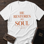 He Restores My Soul t-shirt in White — Psalm 23:3 Christian tee flat lay with Holy Bible and ichthys on dark wood