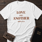 Love One Another t-shirt in White — John 13:34 Christian tee flat lay with Holy Bible and ichthys on dark wood