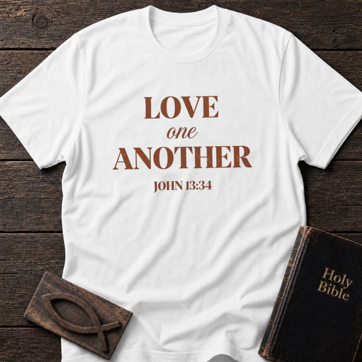 Love One Another t-shirt in White — John 13:34 Christian tee flat lay with Holy Bible and ichthys on dark wood