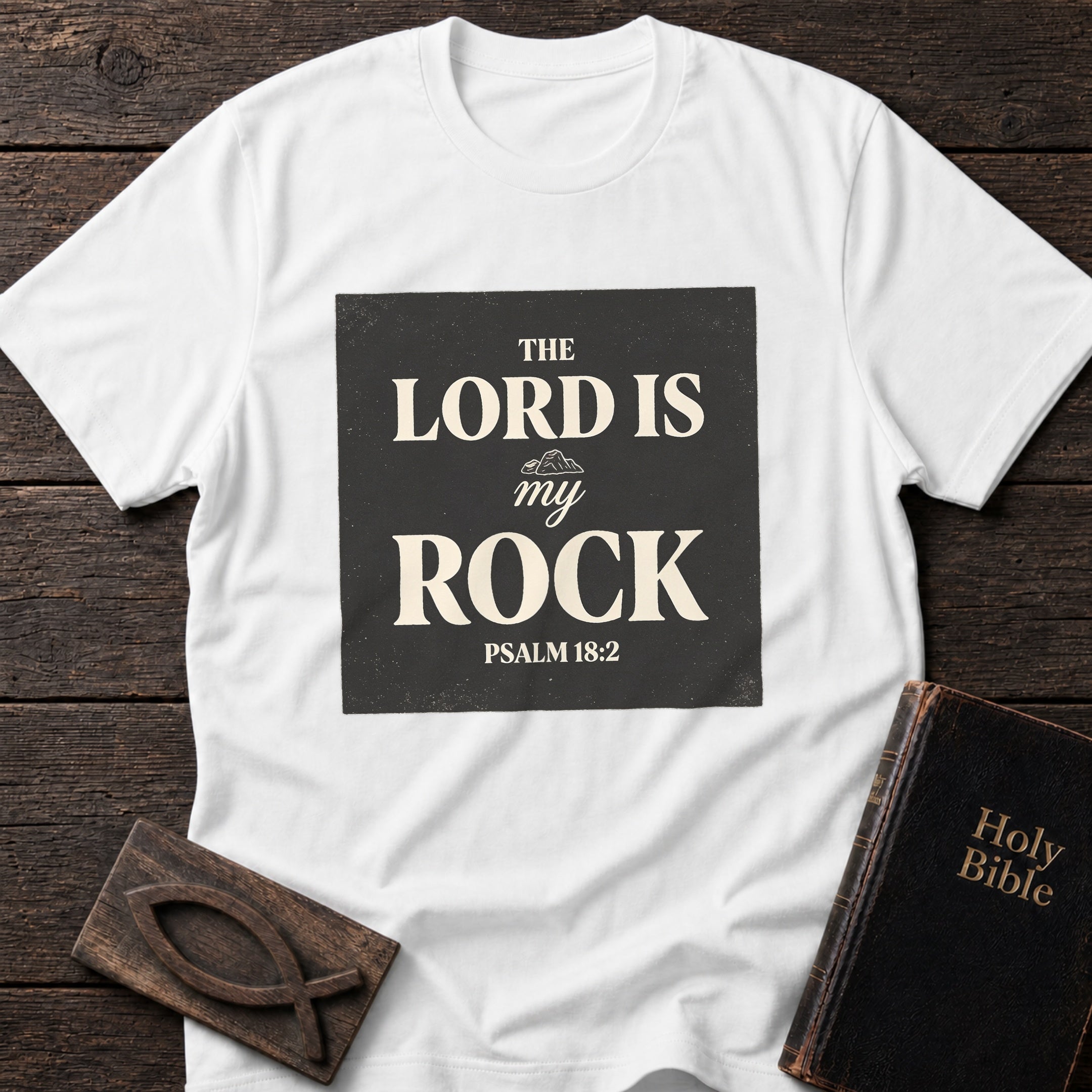The Lord Is My Rock t-shirt in White — Psalm 18:2 Christian tee flat lay with Holy Bible and ichthys on dark wood