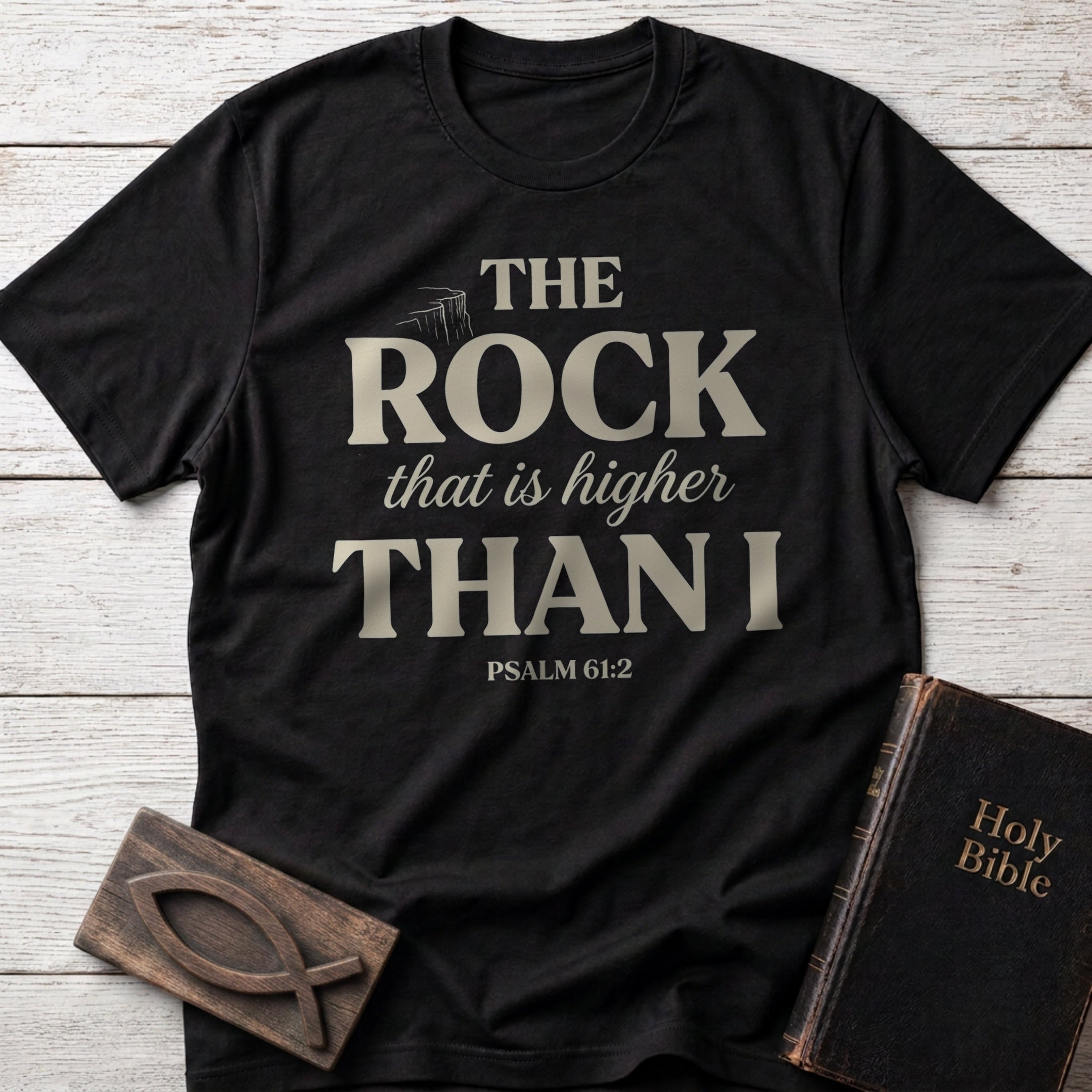 The Rock That Is Higher Than I t-shirt in Black — Psalm 61:2 Christian tee flat lay with Holy Bible and ichthys on whitewashed wood