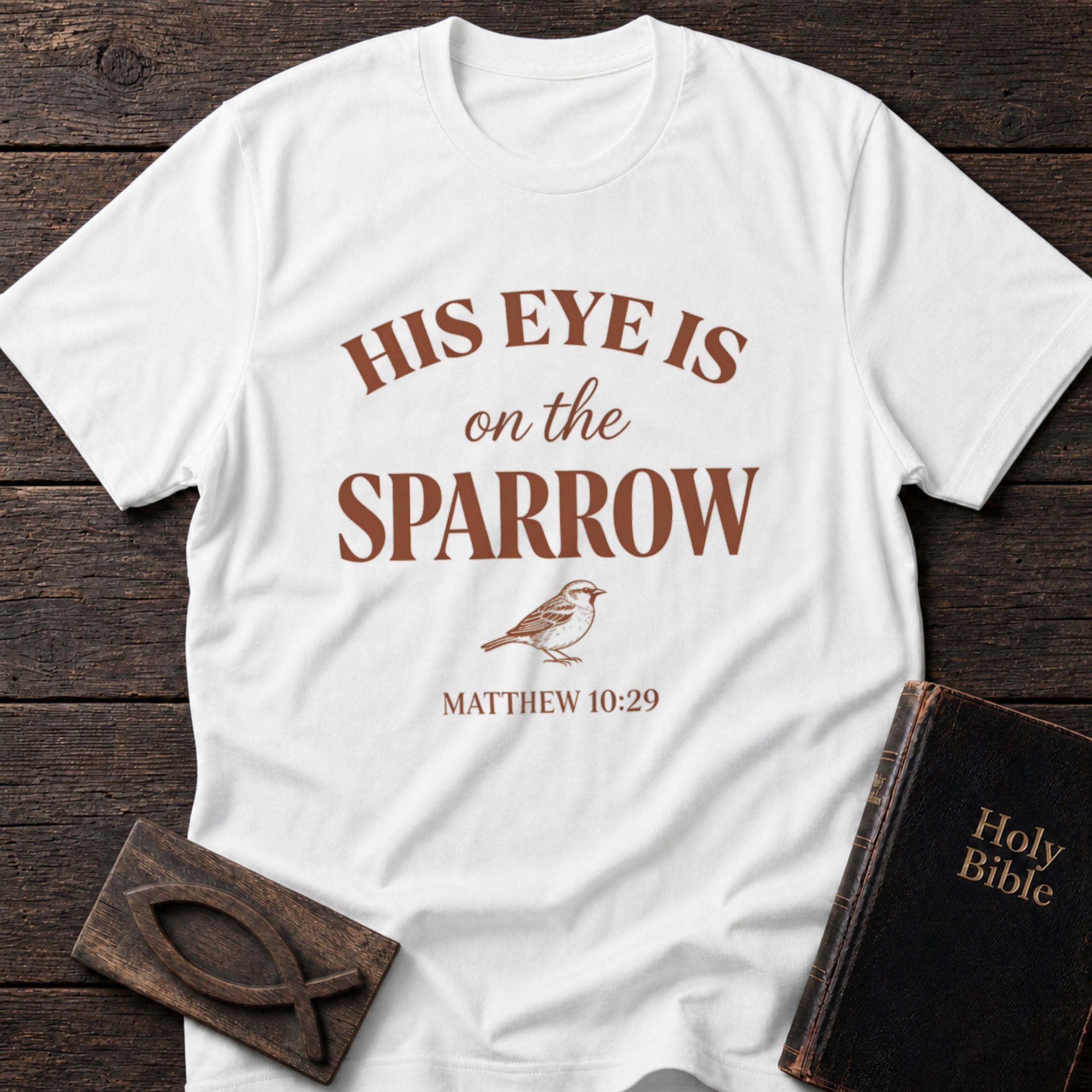 His Eye Is on the Sparrow t-shirt in White — Christian tee flat lay with Holy Bible and ichthys on dark wood
