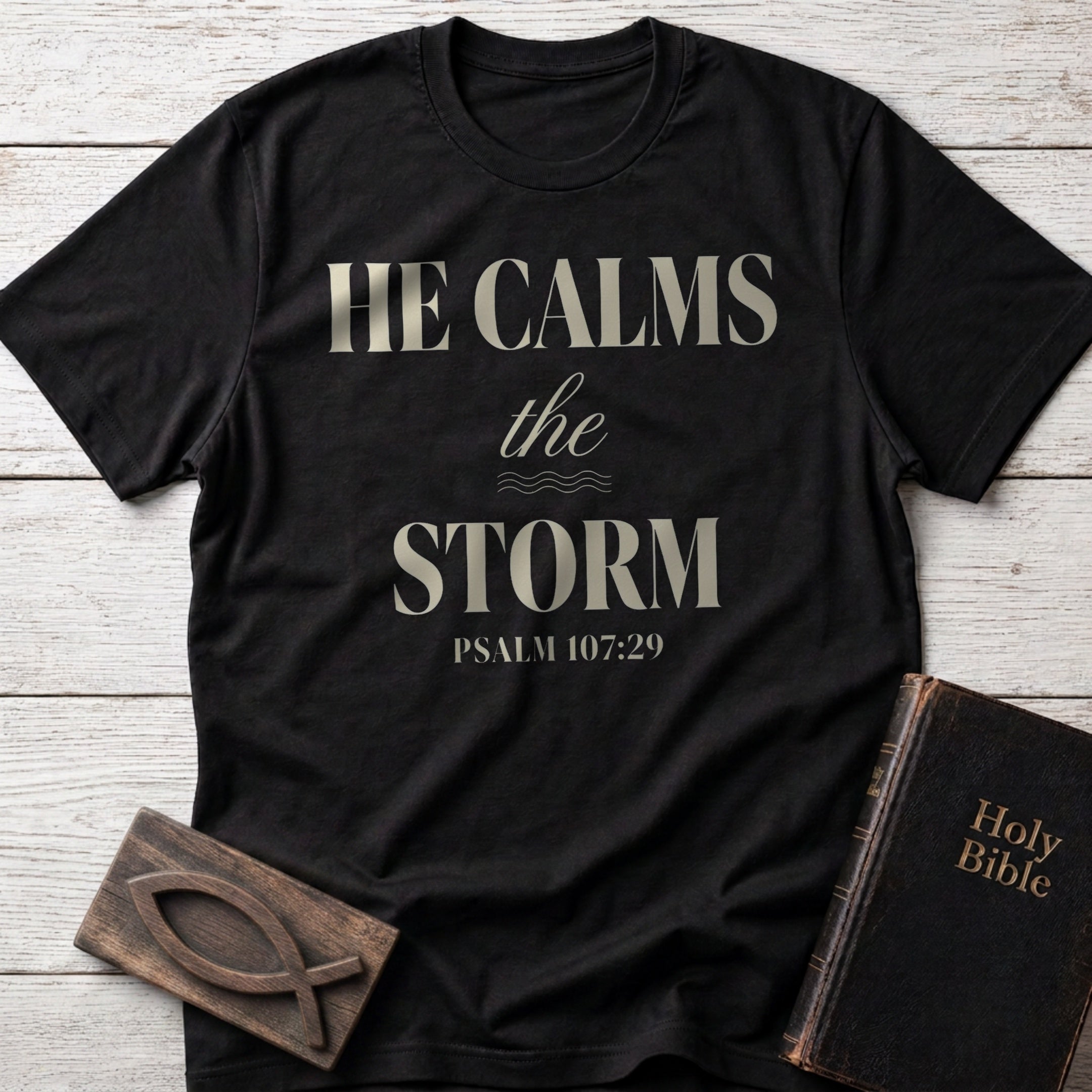 He Calms the Storm t-shirt in Black — Psalm 107:29 Christian tee flat lay with Holy Bible and ichthys on whitewashed wood