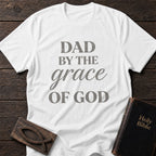 Dad by the Grace of God t-shirt in White — Christian tee flat lay with Holy Bible and ichthys on dark wood