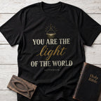 You Are the Light of the World t-shirt in Black — Matthew 5:14 Christian tee flat lay with Holy Bible and ichthys on whitewashed wood