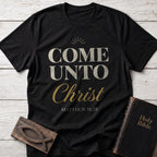 Come Unto Christ t-shirt in Black — Matthew 11:28 Christian tee flat lay with Holy Bible and ichthys on whitewashed wood