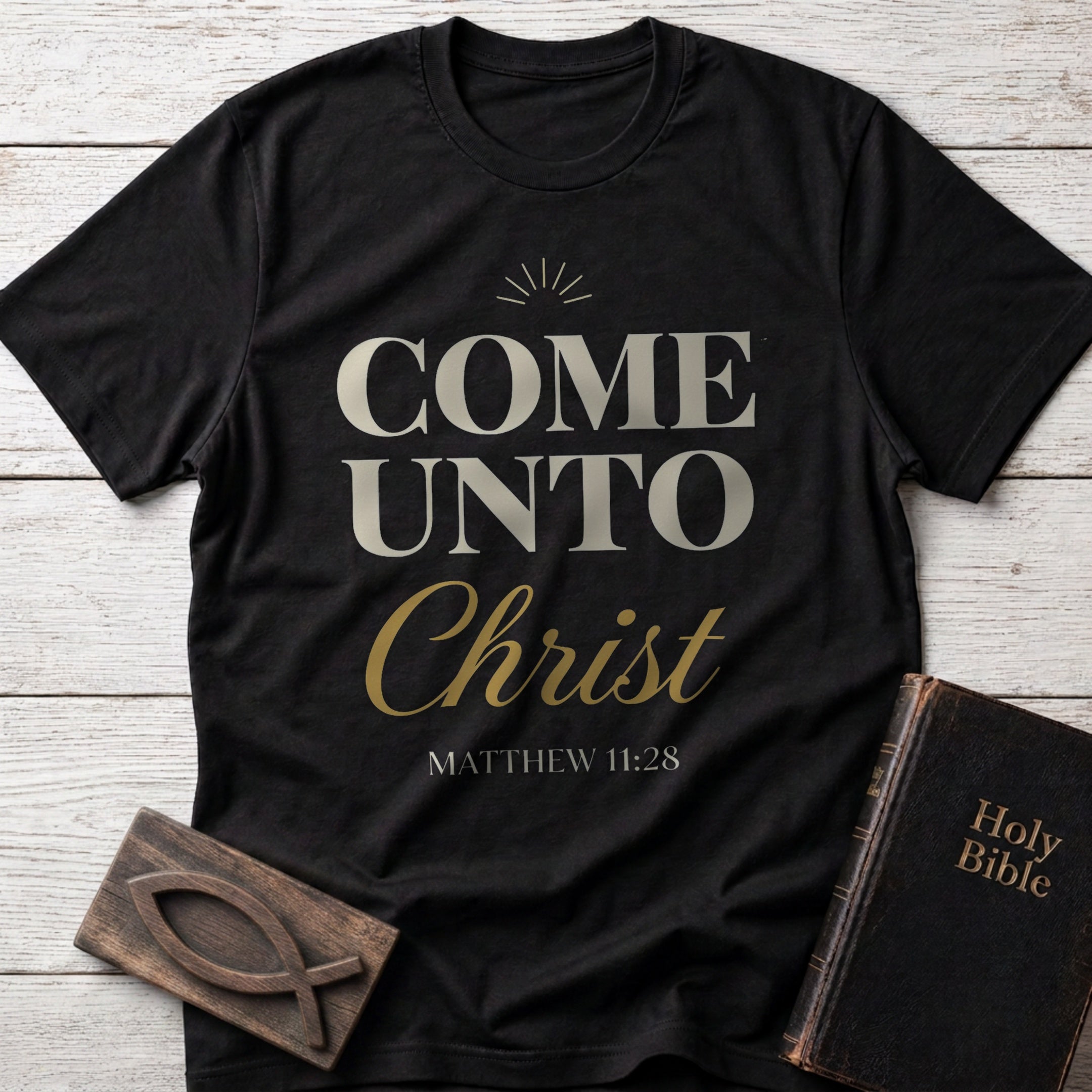 Come Unto Christ t-shirt in Black — Matthew 11:28 Christian tee flat lay with Holy Bible and ichthys on whitewashed wood
