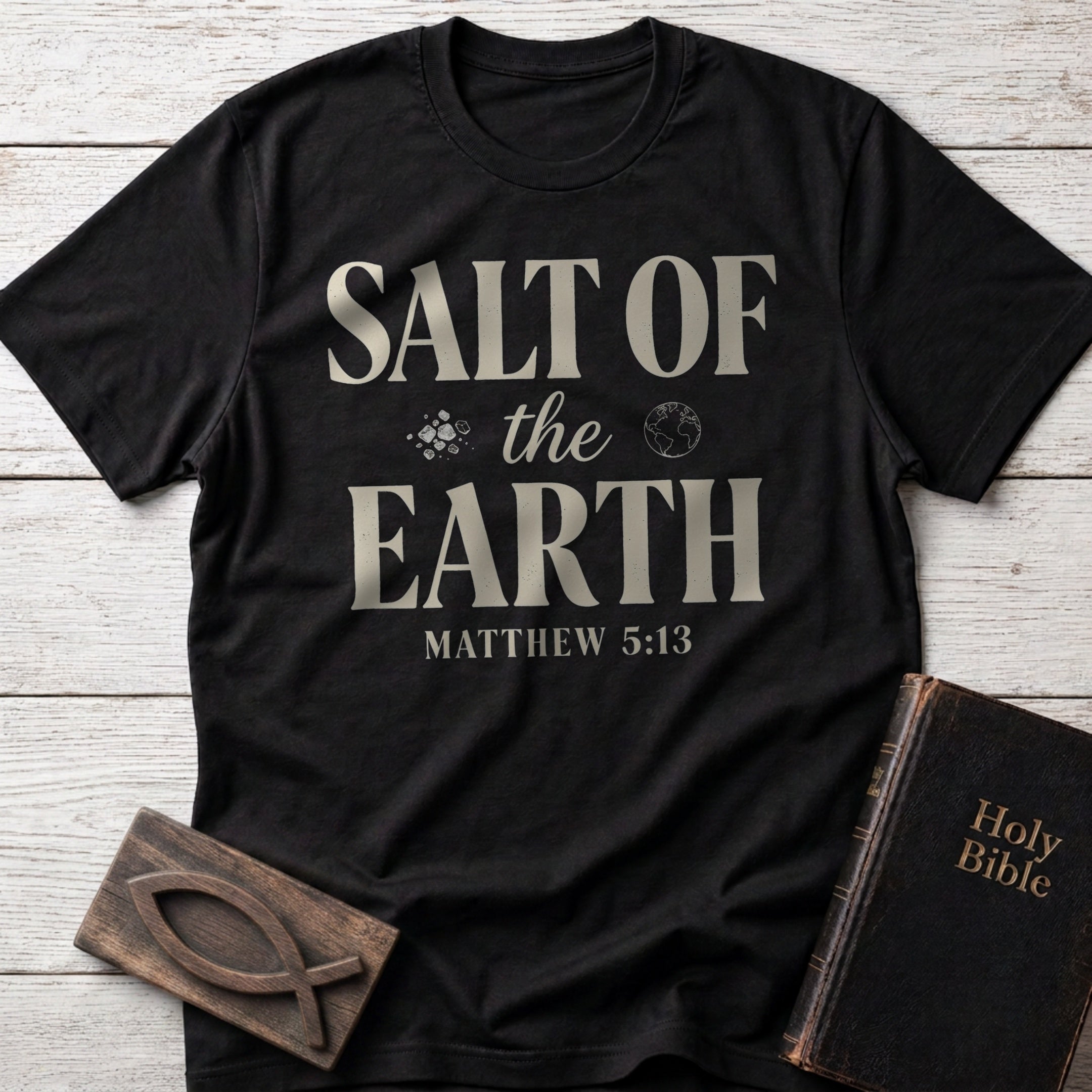 Salt of the Earth t-shirt in Black — Matthew 5:13 Christian tee flat lay with Holy Bible and ichthys on whitewashed wood