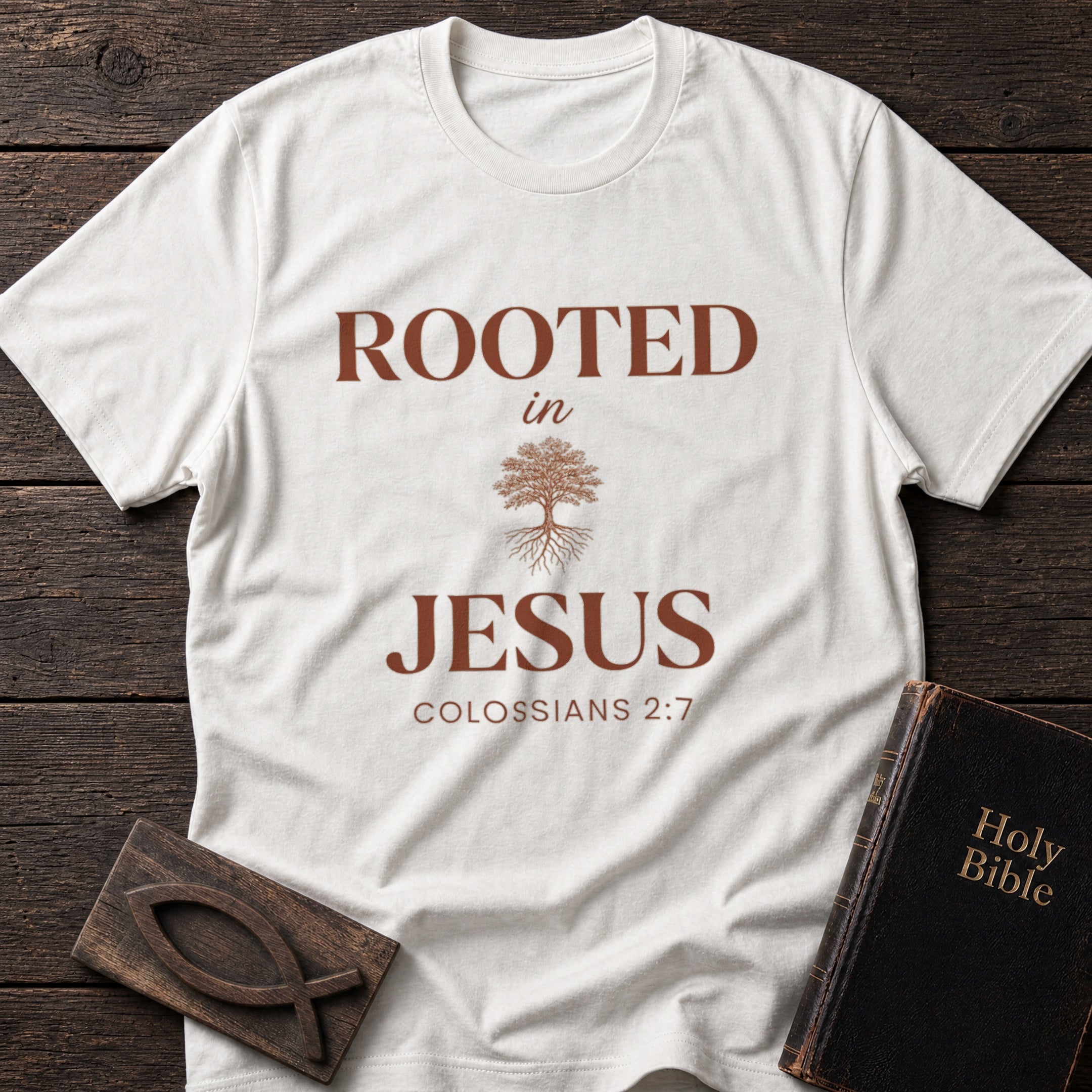 Rooted in Jesus t-shirt in White — Colossians 2:7 Christian tee flat lay with Holy Bible and ichthys on dark wood