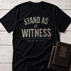 Stand as a Witness t-shirt in Black — Mosiah 18:9 Christian tee flat lay with scripture books on whitewashed wood