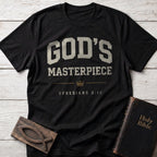 God's Masterpiece t-shirt in Black — Ephesians 2:10 Christian tee flat lay with Holy Bible and ichthys on whitewashed wood