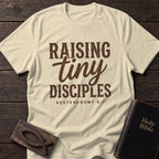 Raising Tiny Disciples t-shirt in Natural — motherhood Christian tee flat lay with Holy Bible and ichthys on dark wood